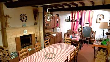 A cozy dining area with a checkered tablecloth, wooden chairs, a fireplace, and a clock on the wall.