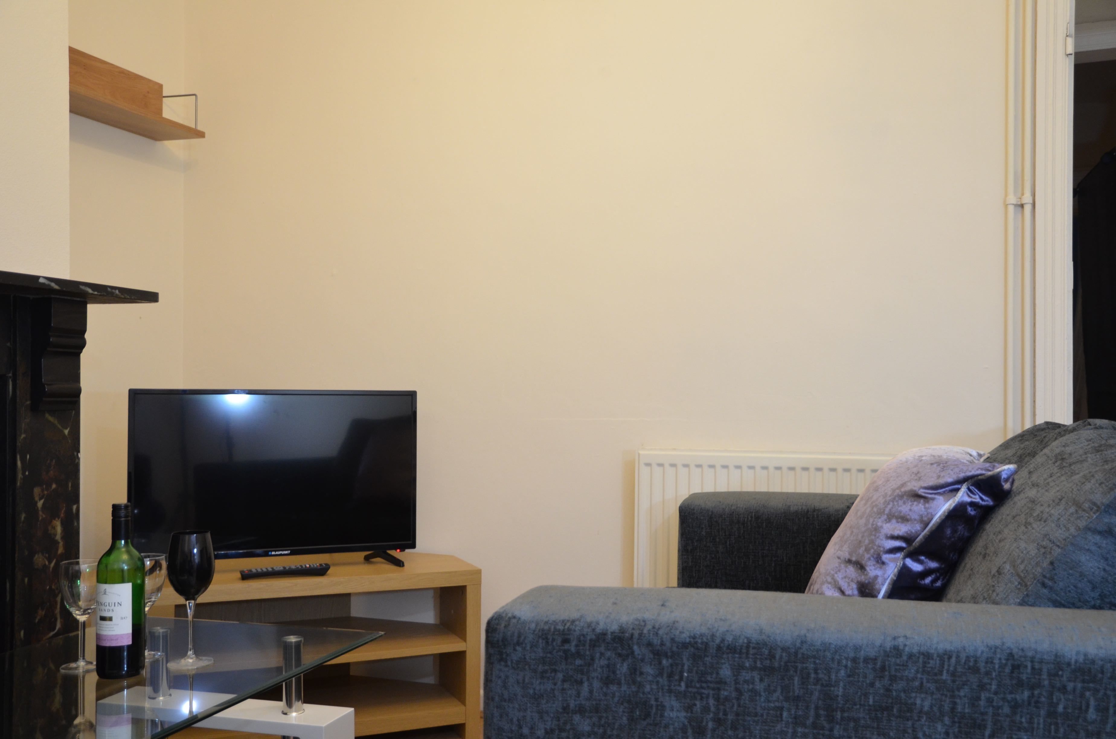 A living room with a flat-screen TV on a wooden stand, a glass table with a wine bottle and glasses, and a grey sofa with a cushion.