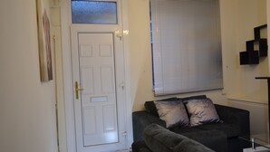 A living room with a white door, a grey sofa with two cushions, a window with blinds, and a small black shelf. - People Boxes Apartments - 38 Ridgway (Luton)