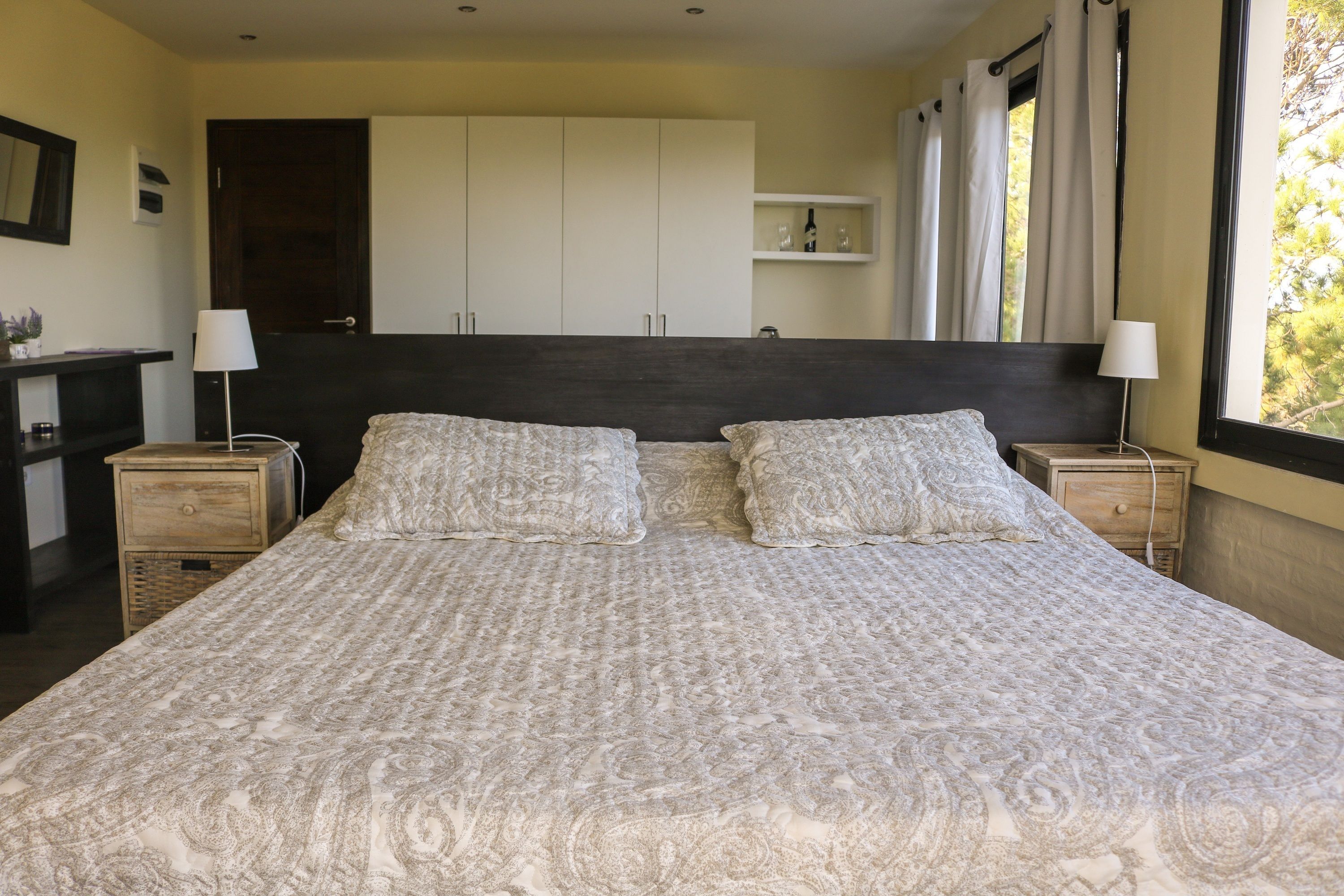 A large bed with a patterned bedspread, two bedside tables with lamps, a wardrobe, and a window with curtains.