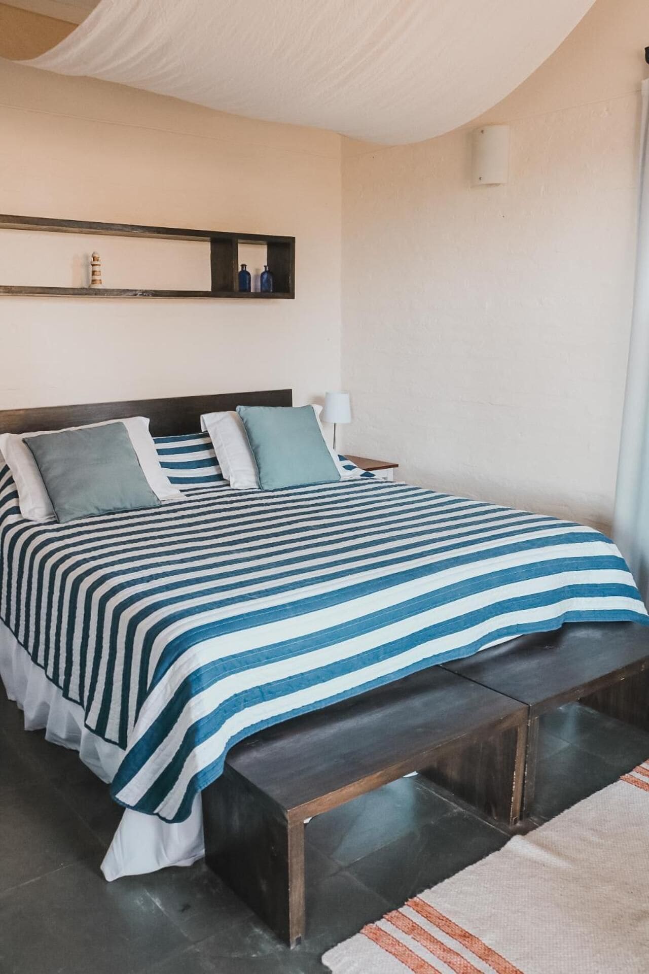 A bedroom with a bed, pillows, a bedside table, a lamp, and a wall-mounted shelf with bottles.