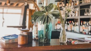 A bar counter with various items including a stack of magazines, a wooden mug, and two vases with plants and flowers. - Posada Azul Marino (José Ignacio)