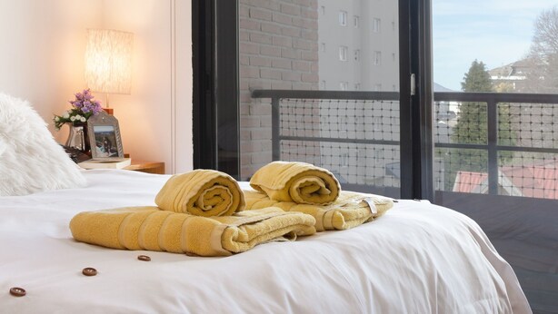 Ein modernes Hotelzimmer mit einem großen Bett, einer Hängelampe, einem Fenster mit Blick auf Gebäude und einem kleinen Tisch mit Lampe und Bilderrahmen.