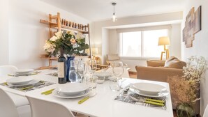 Ein Essbereich mit einem weißen Tisch, gedeckt für vier Personen, umgeben von weißen Stühlen. Eine Holzregal mit Flaschen und eine Blumenvase sind zu sehen. Im Hintergrund sind eine Lampe und ein Sofa erkennbar.