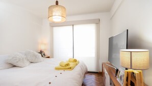 Ein ordentlich bezogenes Bett mit weißer Bettwäsche und aufgerollten gelben Handtüchern, ein Nachttisch aus Holz mit einer Lampe, ein Flachbildfernseher und ein großes Fenster mit vertikalen Jalousien.