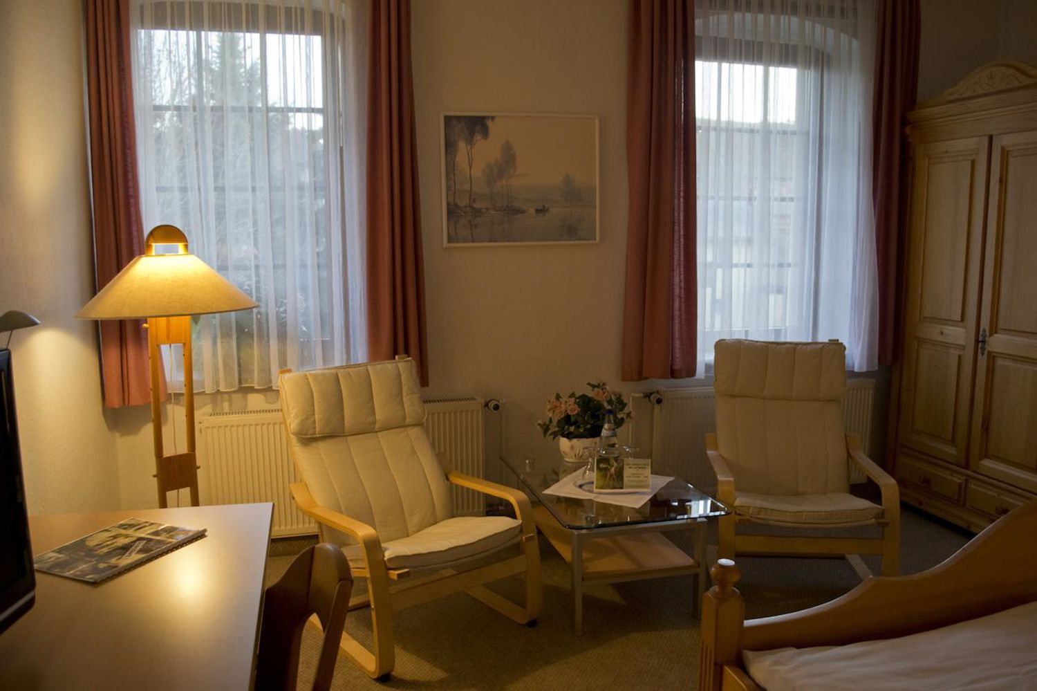 Habitación de hotel con dos sillones, una mesita con un florero, una lámpara, una cama y un radiador.