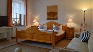 A bedroom with a wooden bed, two bedside tables with lamps, a desk with a television, a window with curtains, and a painting on the wall. - Landhotel Kertscher-Hof (Nobitz)