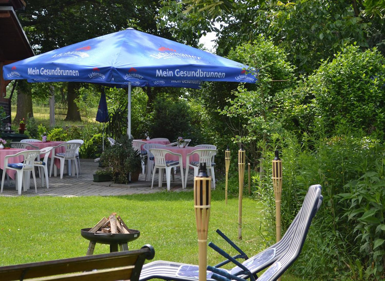 An outdoor seating area with tables and chairs under a blue umbrella, surrounded by greenery.
