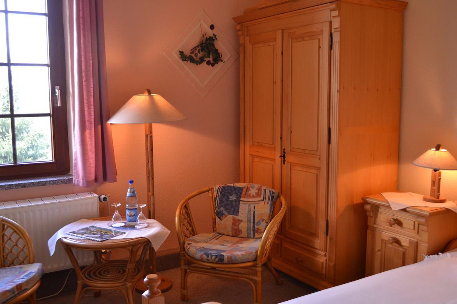 Een knus ingerichte kamer met een houten garderobe, een rieten stoel, een klein tafeltje met een lamp en een raam met gordijnen.