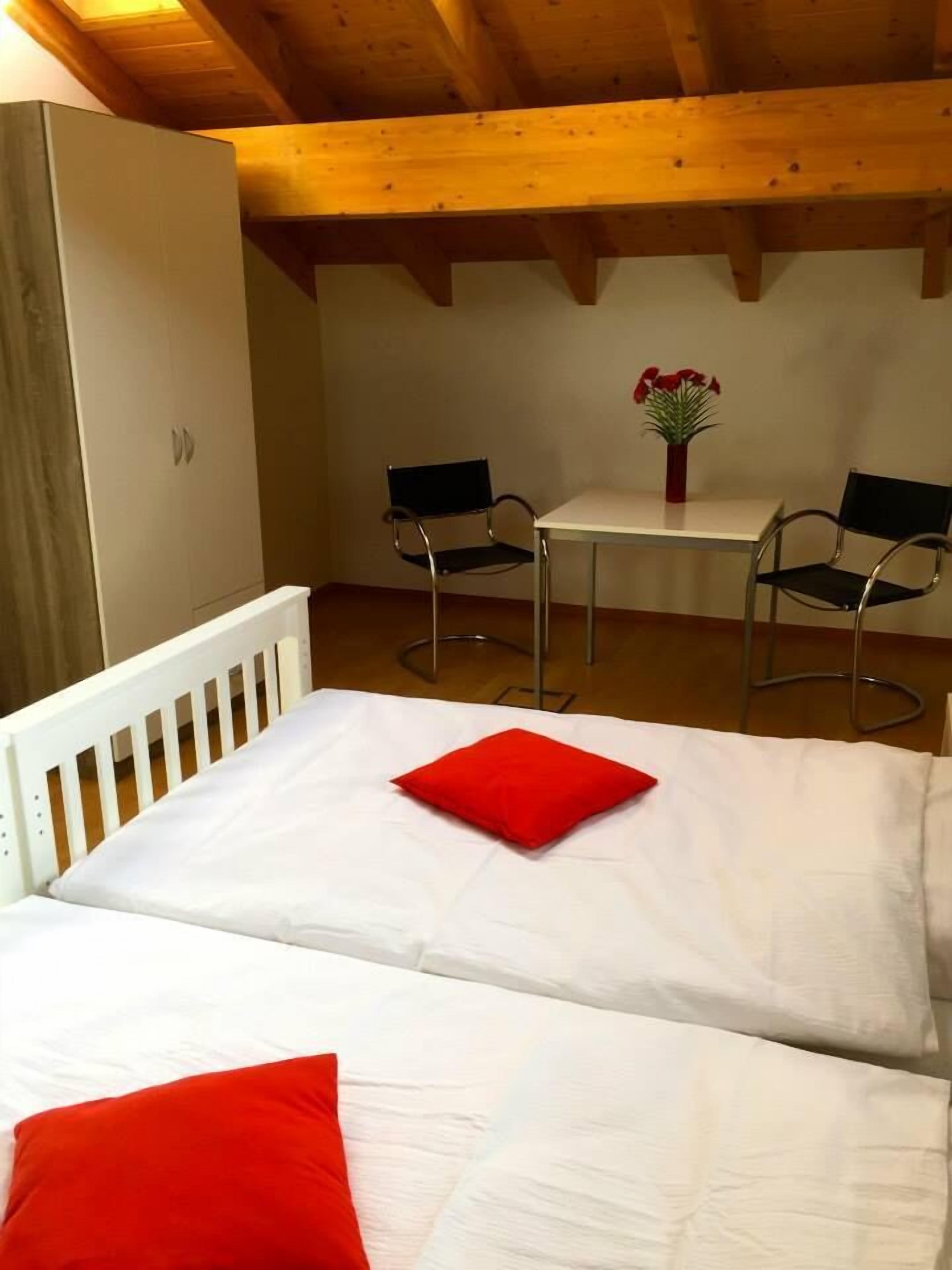Ein Schlafzimmer mit einem weißen Bett, roten Kissen, einem kleinen Tisch mit Stühlen und einer Vase mit Blumen.