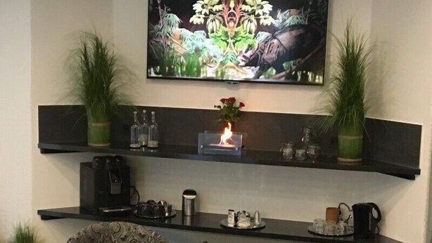 A living room with two patterned armchairs, a coffee table with a vase of flowers, a flat-screen TV mounted above a black shelf with decorative items, and a fireplace with a lit fire.
