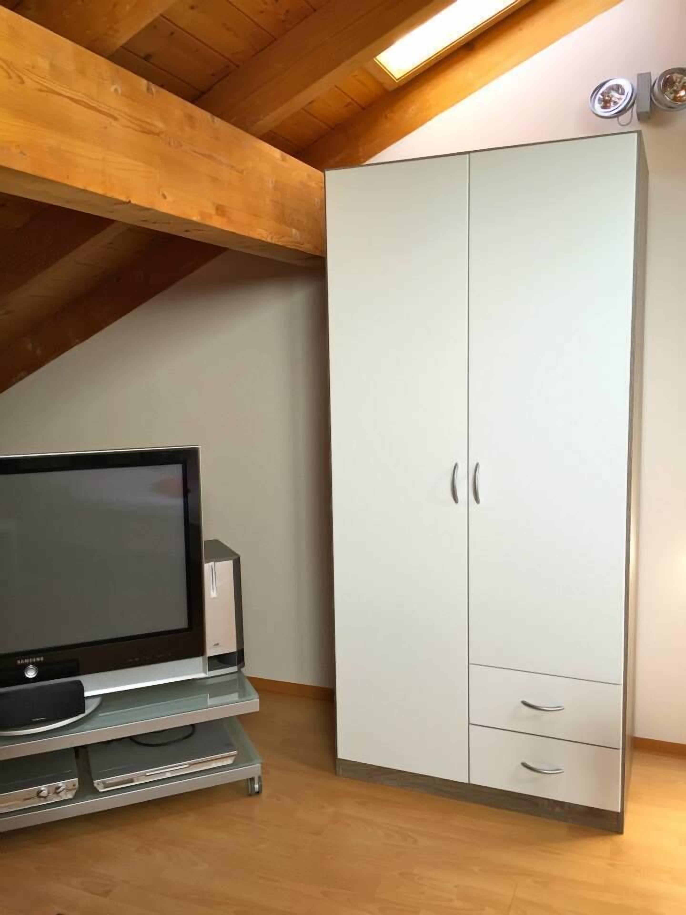 A room with a wooden ceiling, a white wardrobe with two doors and two drawers, and a flat-screen TV on a stand.