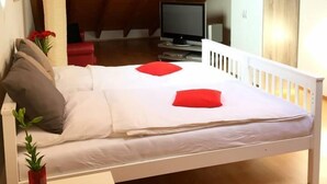 A bedroom with a wooden ceiling, a bed with white bedding and red pillows, a TV, and a small table with a red vase.