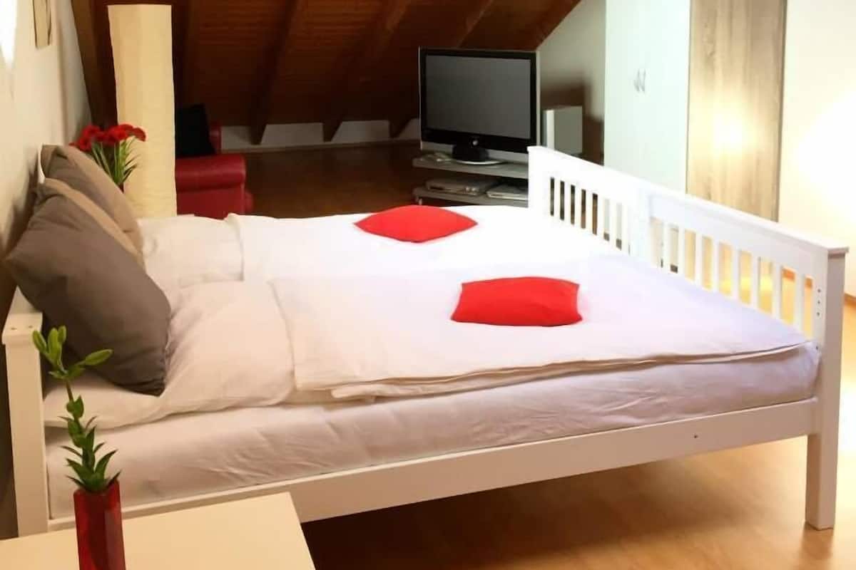 Een slaapkamer met een houten plafond, een bed met witte lakens en rode kussens, een televisie en een klein tafeltje met een rode vaas.