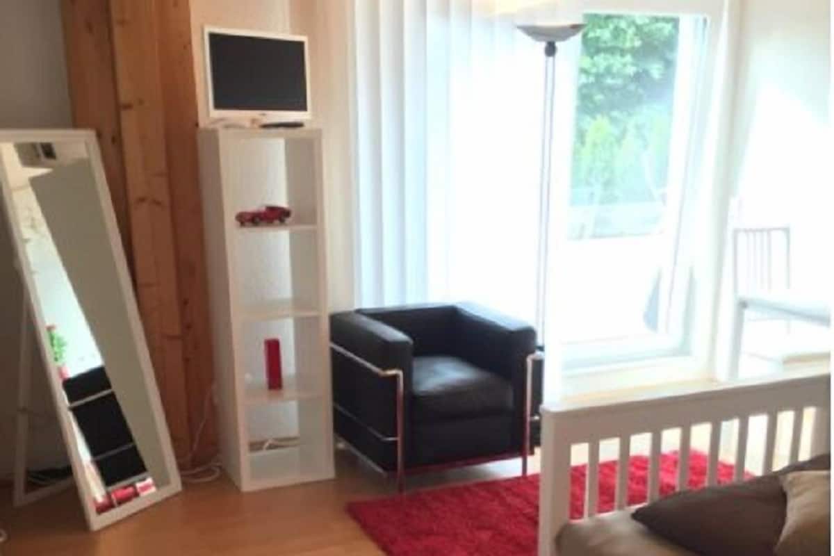 Een kamer met een wit ledikant, een rood vloerkleed, een zwarte stoel en een witte kast met een televisie.