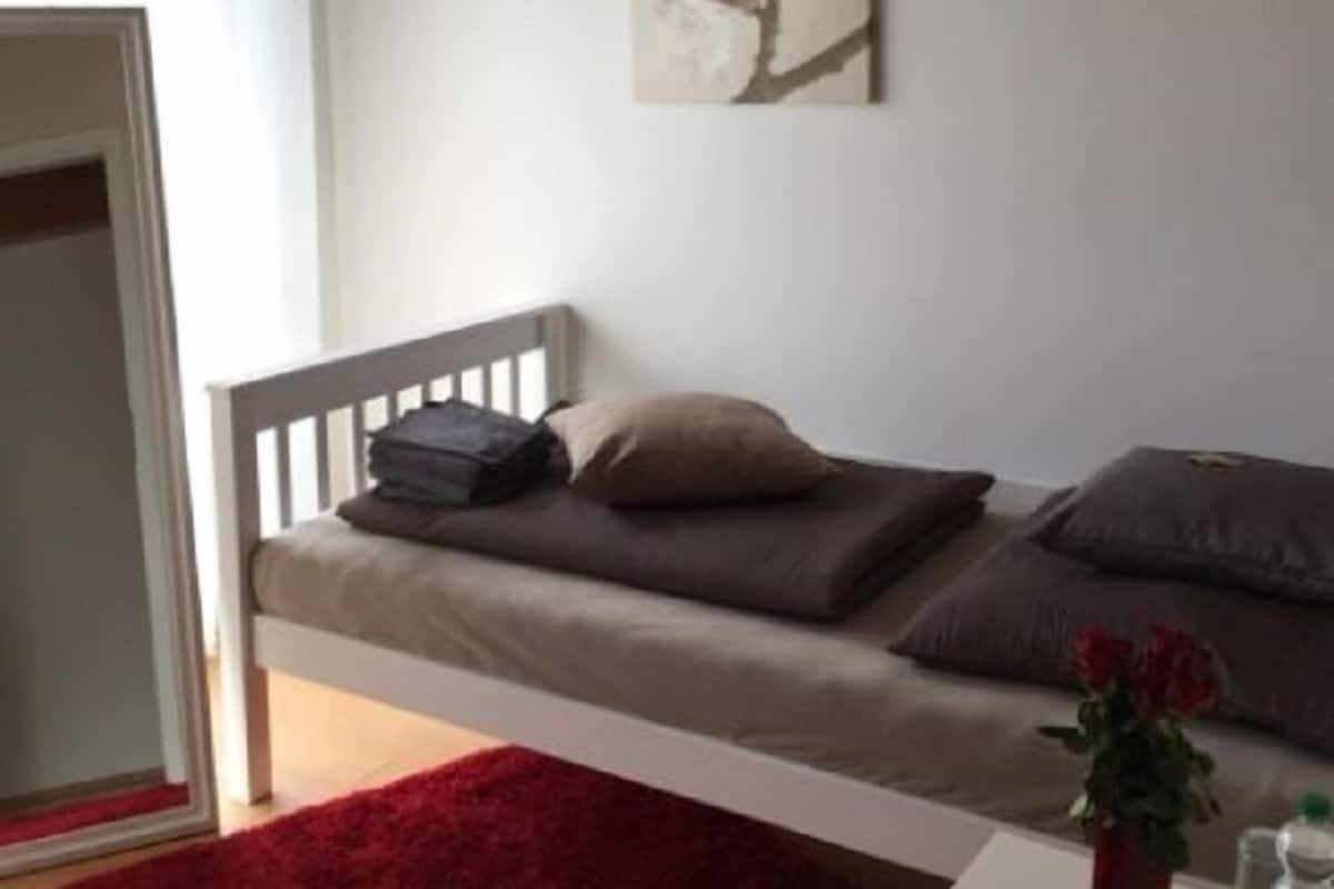 Een slaapkamer met een bed, kussens, een rood vloerkleed, een vaas met bloemen en een schilderij aan de muur.