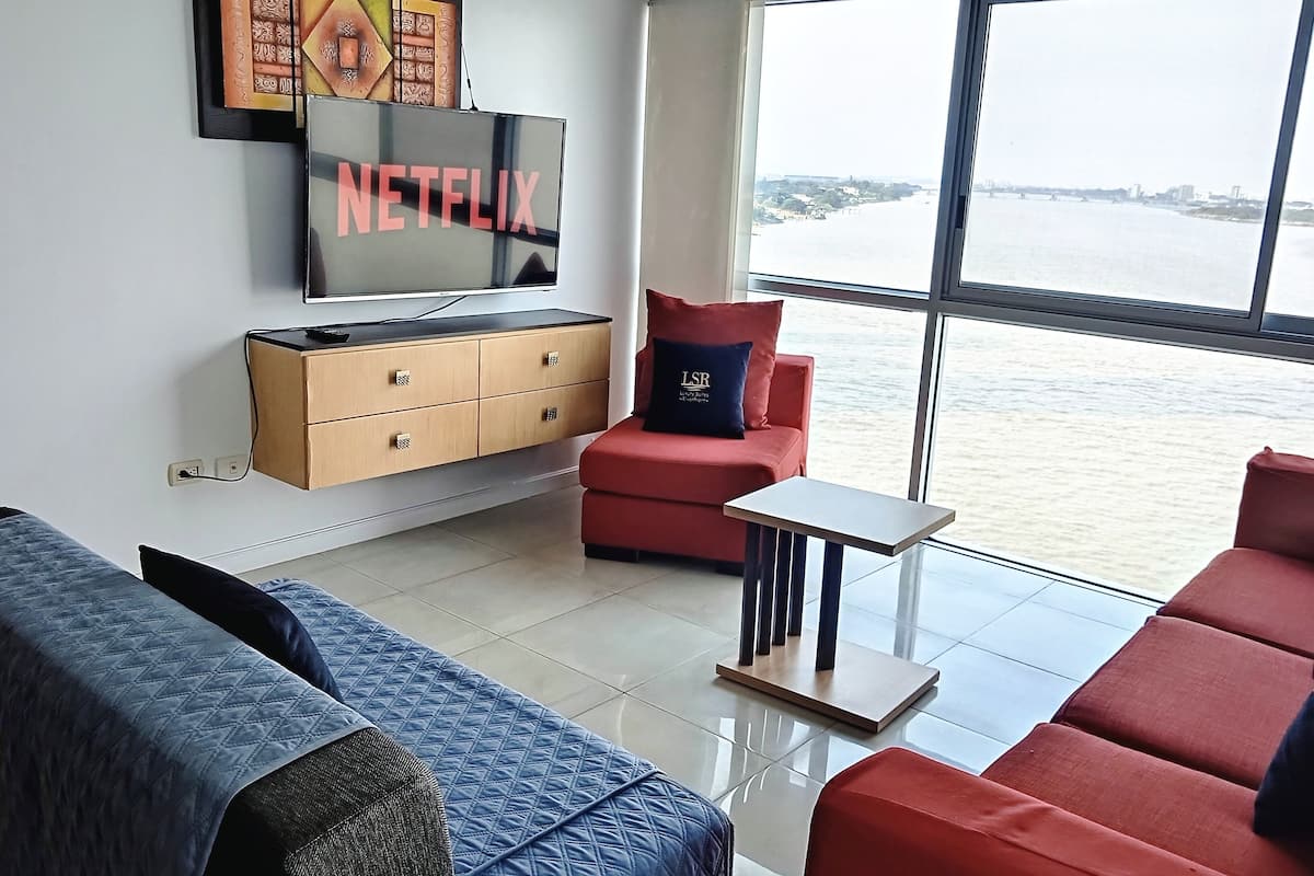 Una sala de estar moderna con un ventanal grande, un sofá rojo, un sofá azul, una mesita pequeña y un televisor con el logotipo de Netflix.