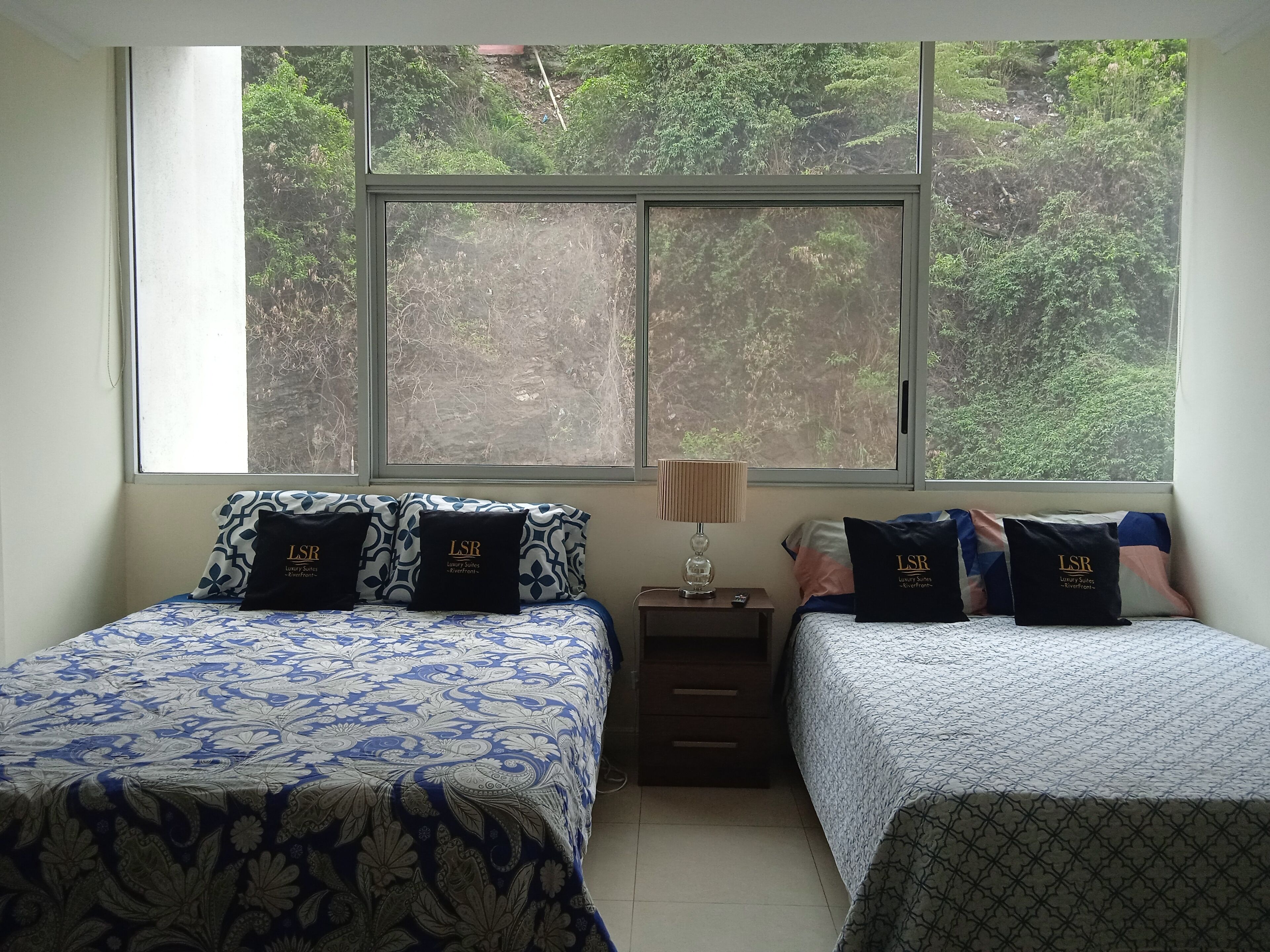 A bedroom with two beds, each with patterned bedding and pillows labeled 'LSR Luxury Suites'. A nightstand with a lamp and remote control is between the beds. Large windows offer a view of greenery outside.