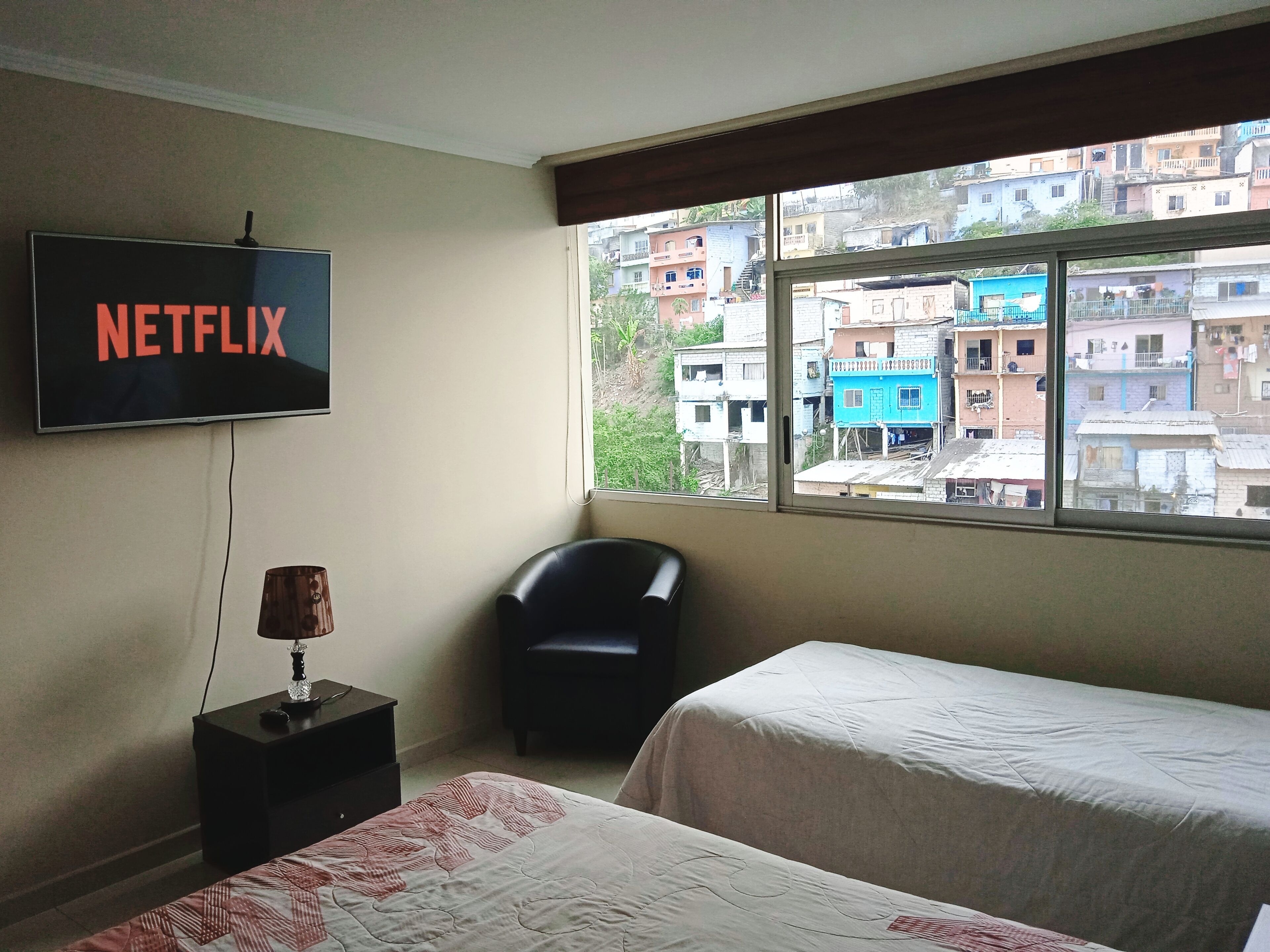 A hotel room with a bed, a chair, a TV displaying 'NETFLIX', and a window with a view of buildings.