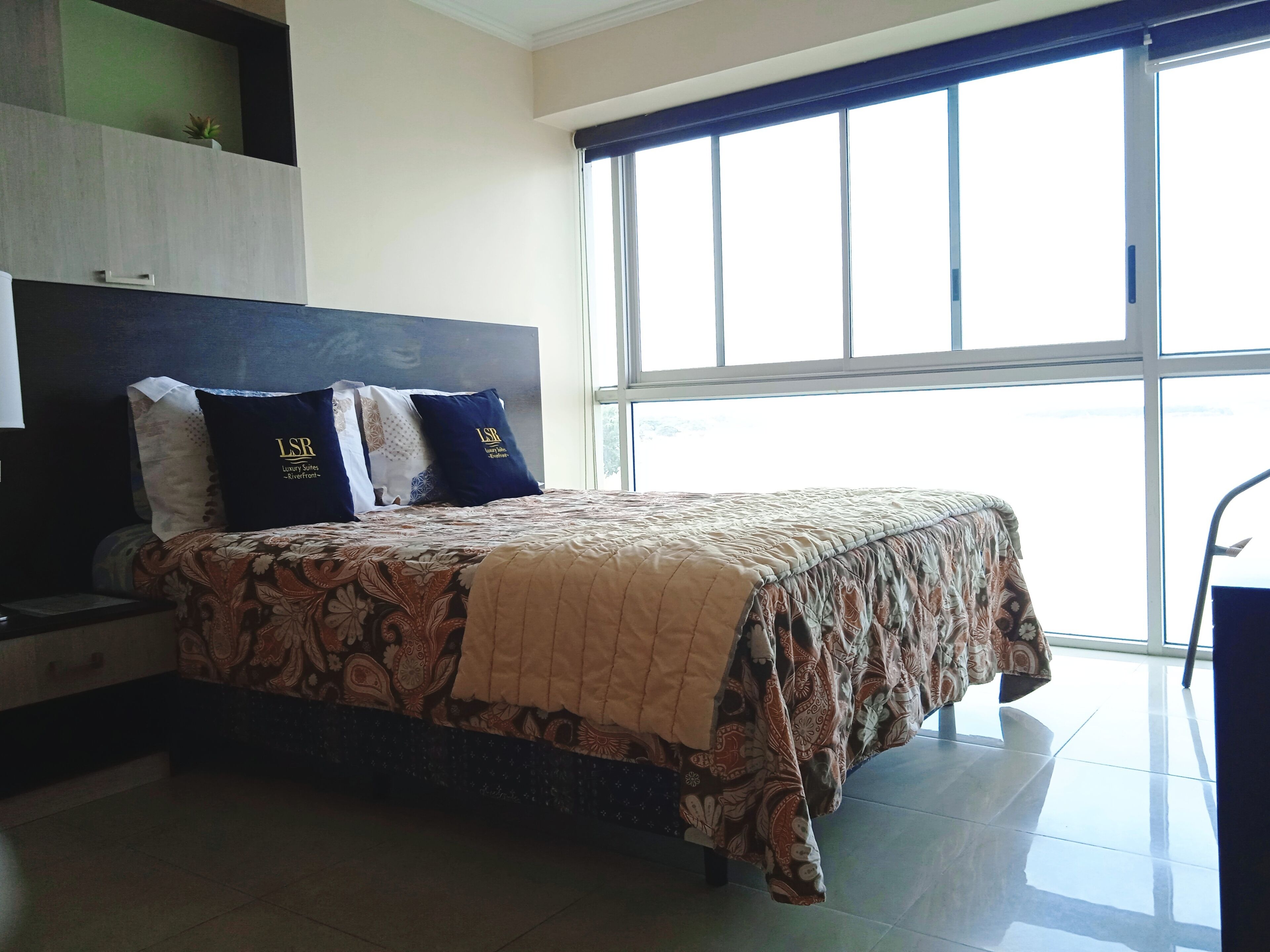 A hotel room with a bed, a nightstand, a dresser, and a large window.