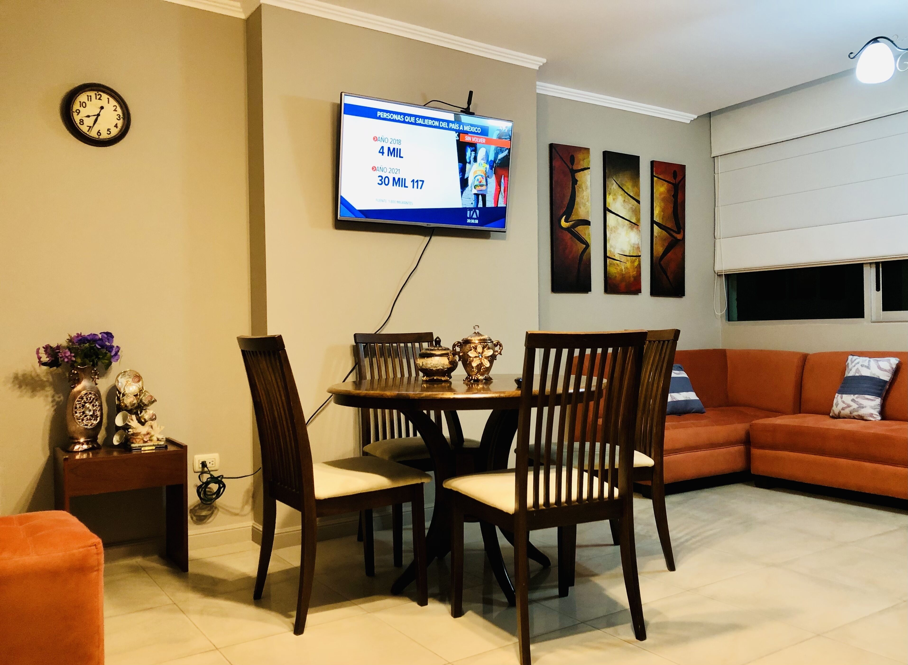 Ein Wohnzimmer mit Esstisch, zwei Stühlen, einer Couch, einem an der Wand befestigten Fernseher und einer Wanduhr.