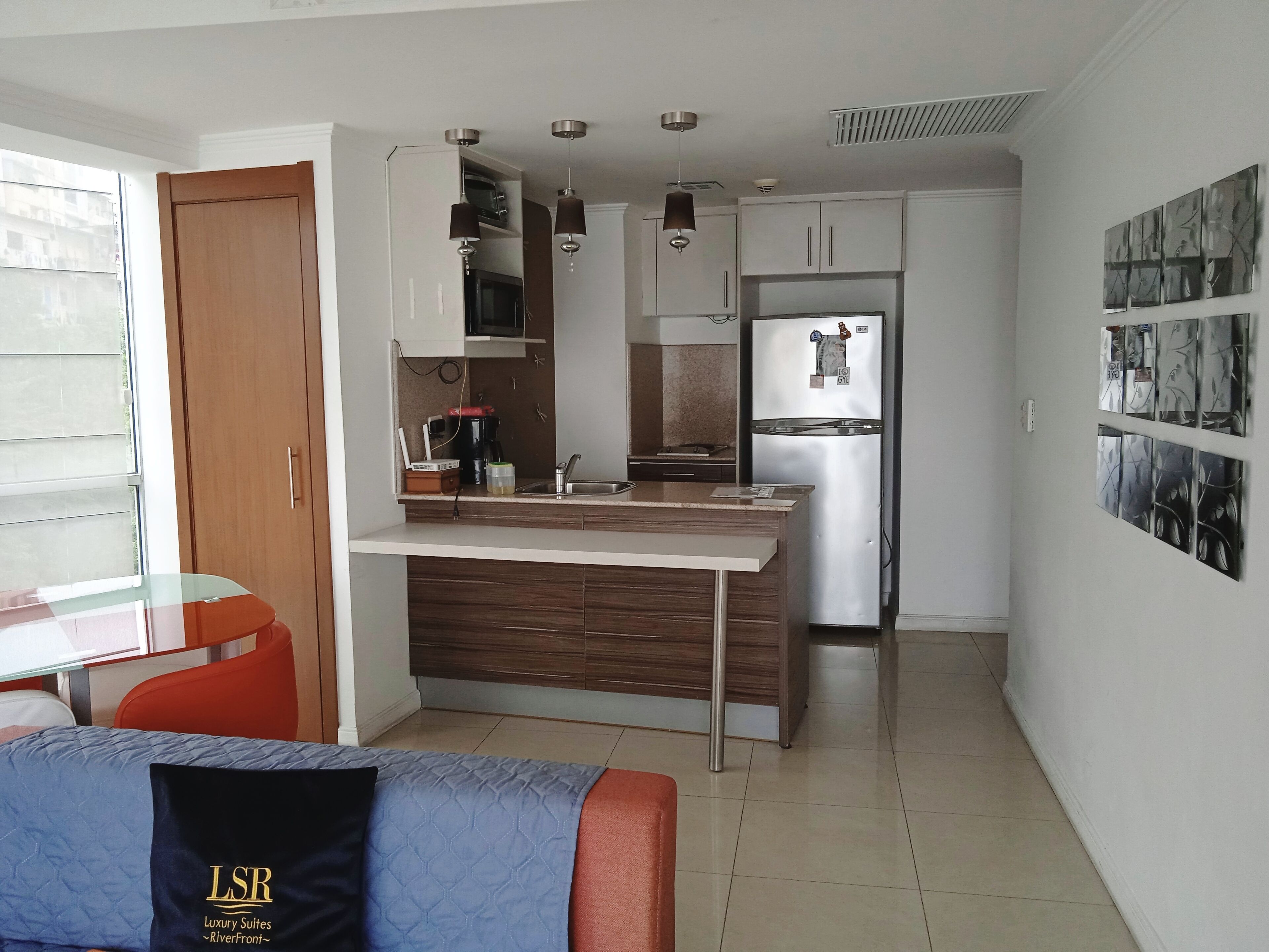 Cocina compacta con microondas integrado, refrigerador y cafetera. Hay un comedor con mesa de vidrio y sofá.