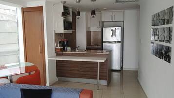 Cocina compacta con microondas integrado, refrigerador y cafetera. Hay un comedor con mesa de vidrio y sofá.