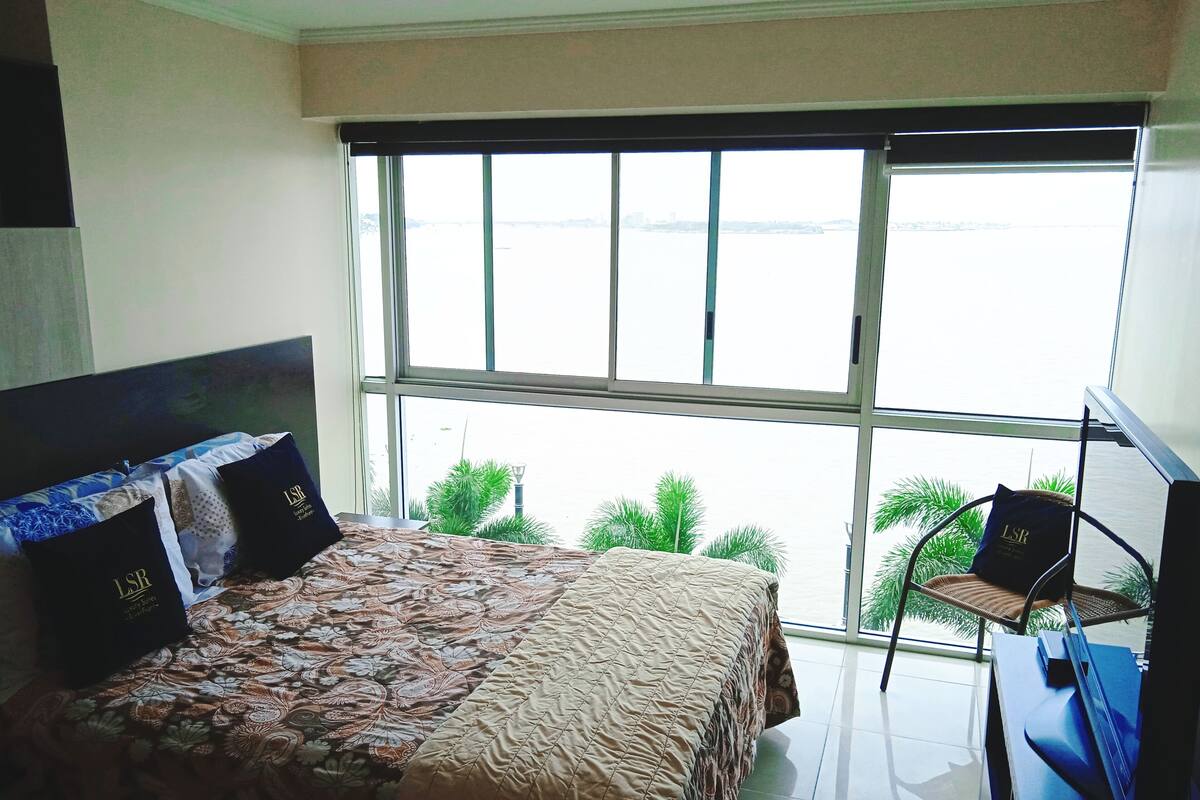 Un dormitorio con una cama grande, una silla y vista a un cuerpo de agua a través de un ventanal amplio.