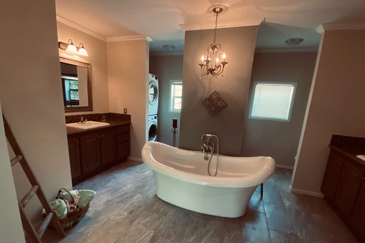 A spacious bathroom with a freestanding bathtub, a double sink vanity, a washing machine, and a chandelier.