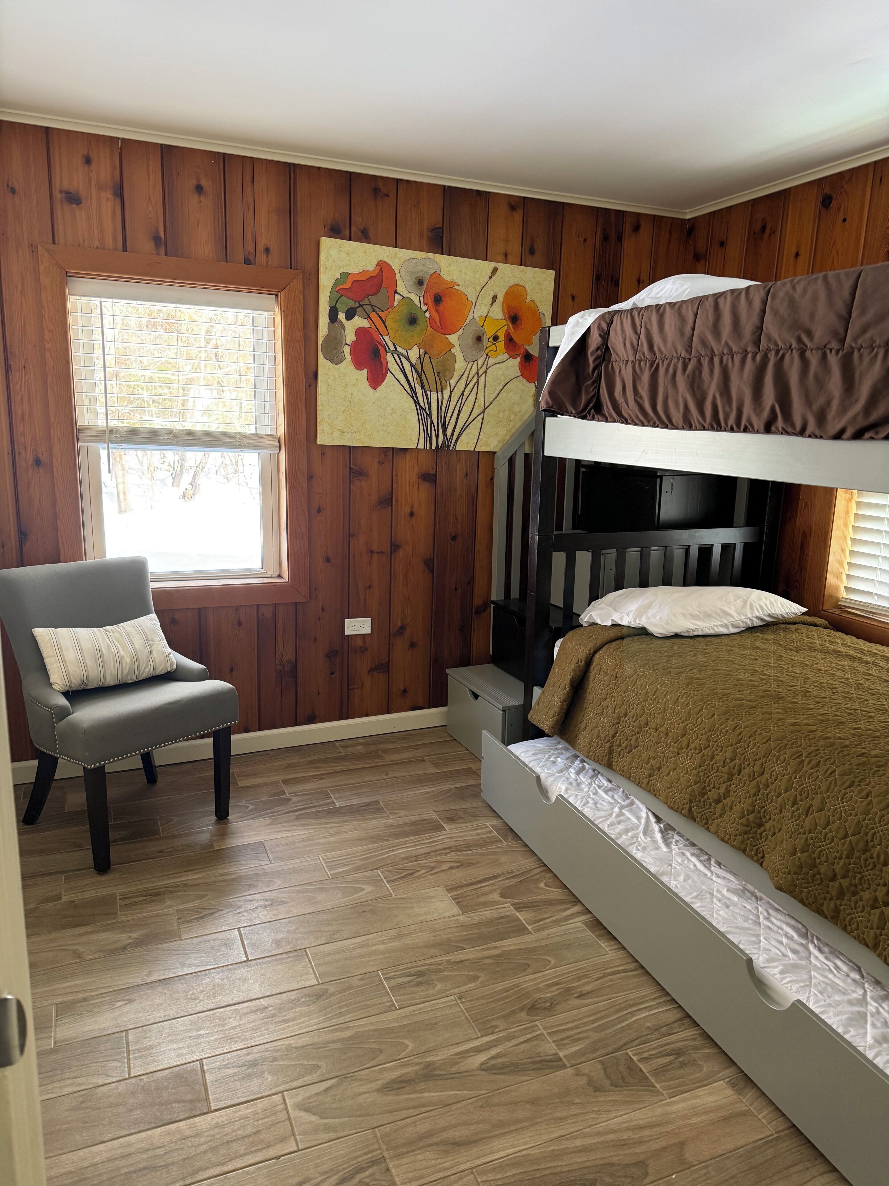 Quarto com beliche, uma cadeira com almofada, piso de madeira, janela com persianas e um quadro na parede.