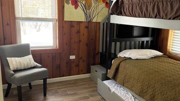 Quarto com beliche, uma cadeira com almofada, piso de madeira, janela com persianas e um quadro na parede.
