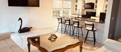 A modern living room with a large sofa, a coffee table, and a swan sculpture. There is a kitchen area with a bar and seating.