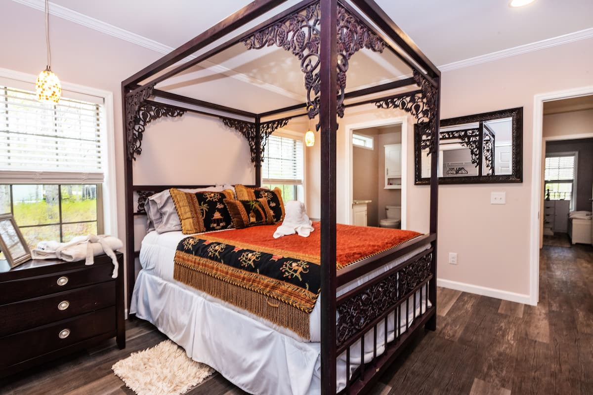 A four-poster bed with a white bedspread and decorative pillows, a wooden nightstand with drawers, and a fluffy rug.