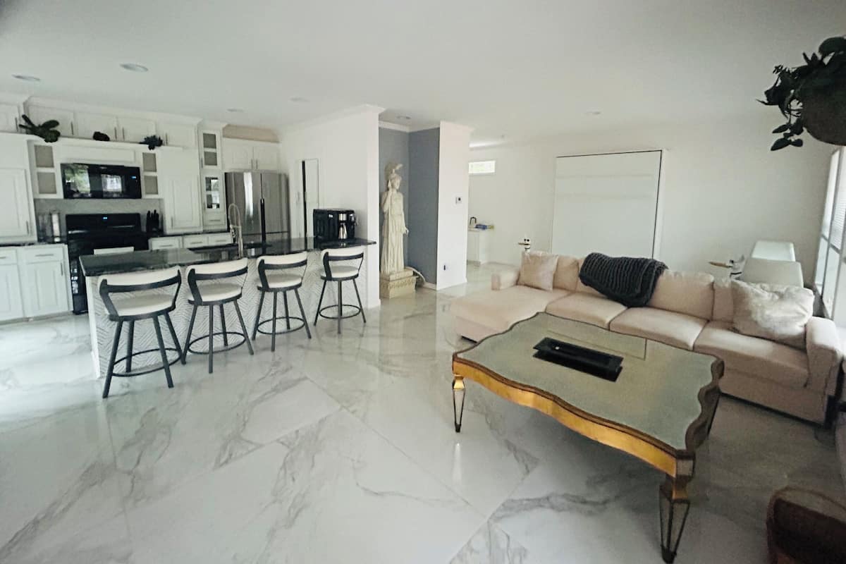 A modern living room with a marble floor, a beige sofa, a coffee table, and a dining area with chairs and a kitchen in the background.