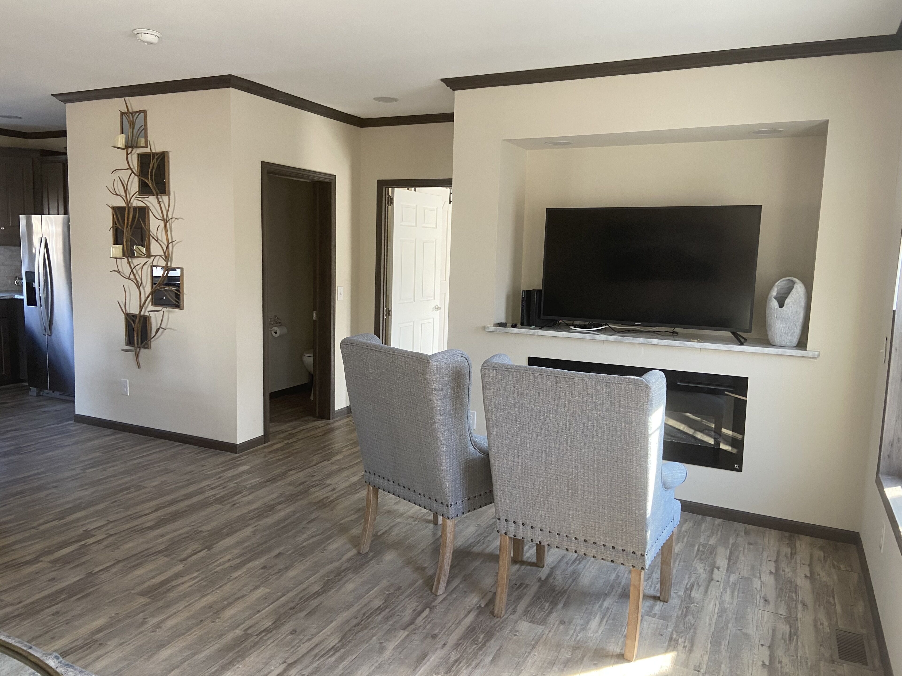 Un salón amplio con una televisión de pantalla plana montada en la pared, dos sillones grises tapizados y suelo de madera.