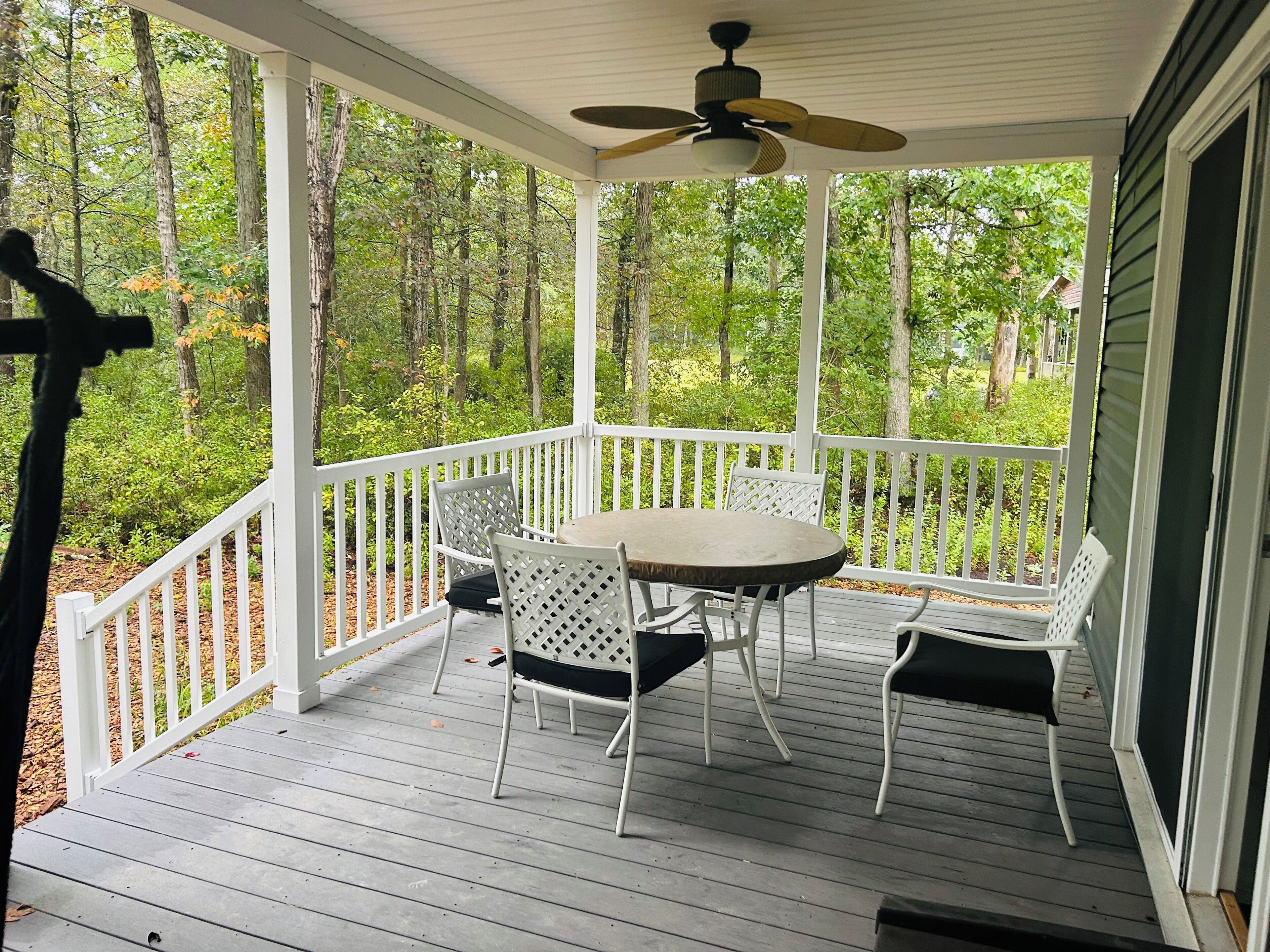 A covered porch with a table and chairs, surrounded by a white railing, overlooking a forested area.