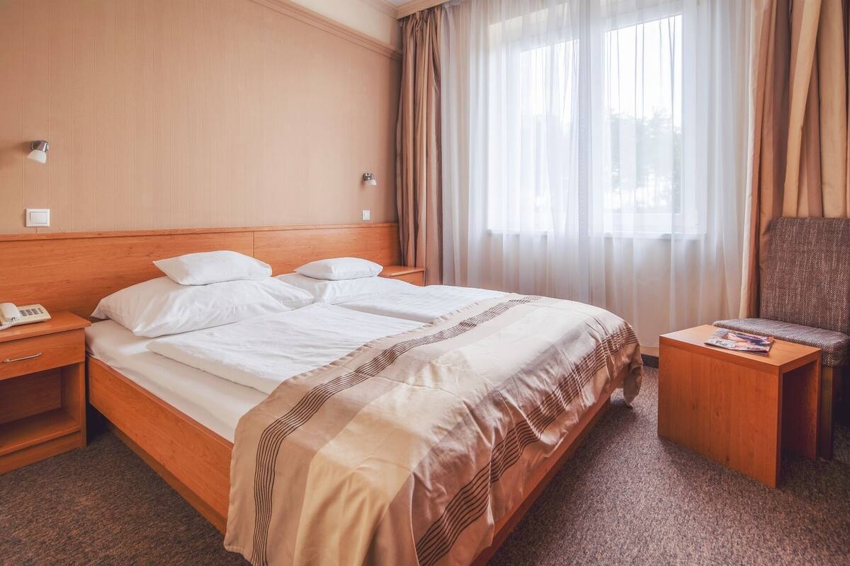 Habitación de hotel con cabecera de madera, cama con ropa blanca, mesita de noche con lámpara, una silla y una pequeña mesa con una revista.