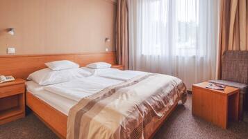 A hotel room with a wooden headboard, a bed with white linens, a bedside table with a lamp, a chair, and a small table with a magazine.