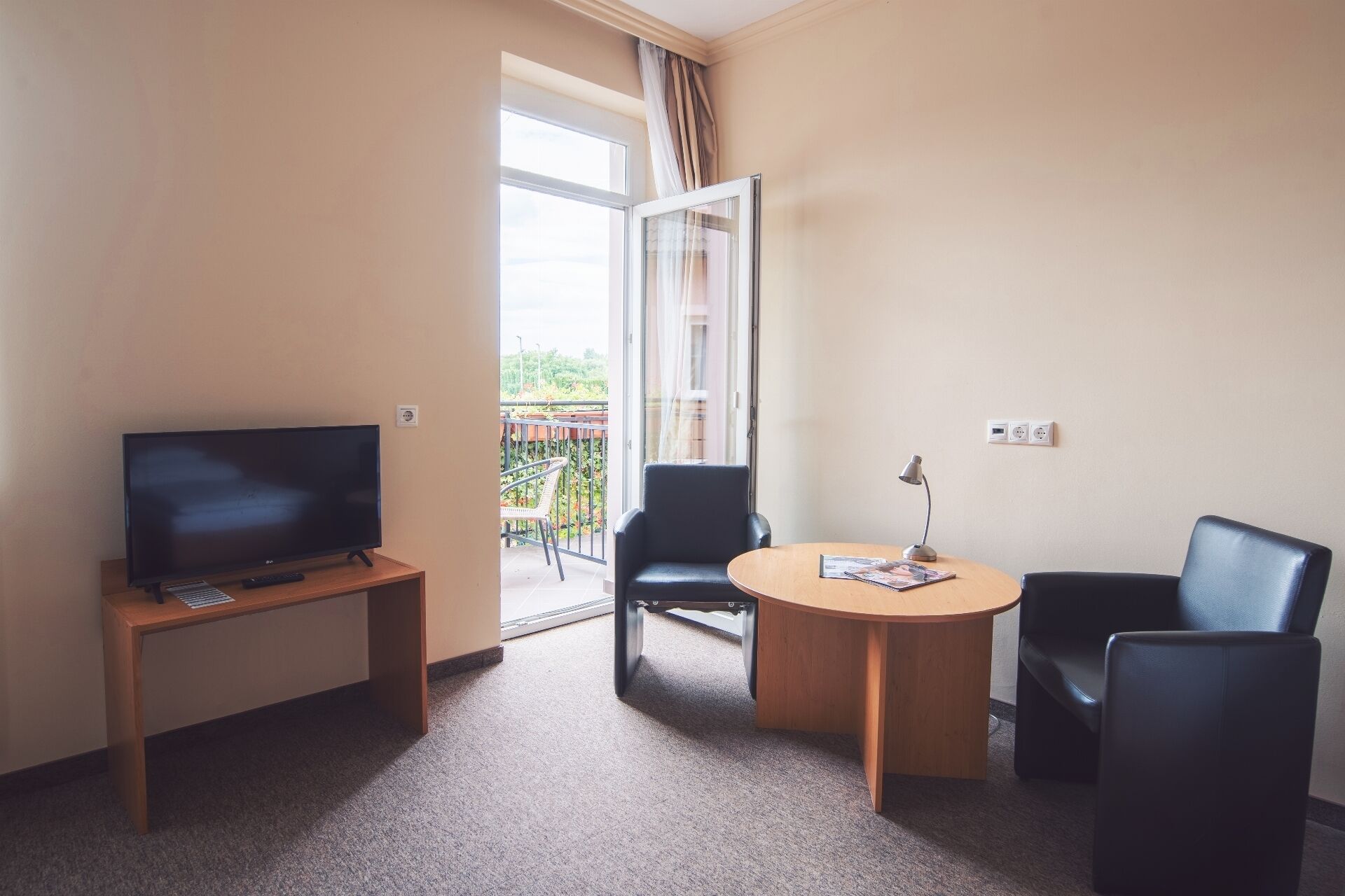 A hotel room with a TV on a wooden stand, a round table with a lamp, two black chairs, and a balcony with a view of greenery.