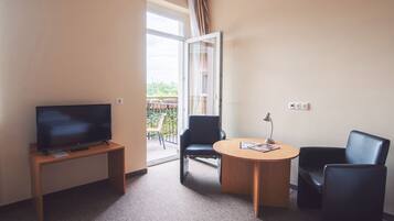 Ein Hotelzimmer mit Fernseher auf einem Holztisch, einem runden Tisch mit Lampe, zwei schwarzen Stühlen und einem Balkon mit Blick auf Grünflächen.