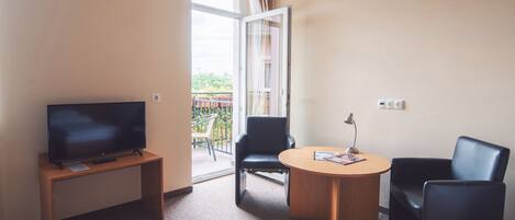 A hotel room with a TV on a wooden stand, a round table with a lamp, two black chairs, and a balcony with a view of greenery.