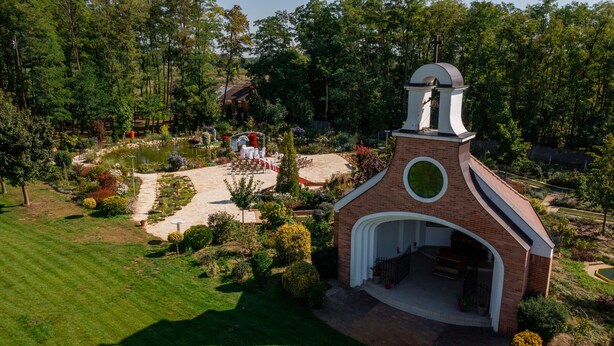 A small brick chapel with a cross on top, surrounded by a garden with a pond, chairs, and a pathway. - Granada Konferencia Wellness Sport Hotel (Kecskemet)