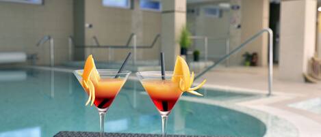 Two cocktails on a table by a poolside with a clear view of the pool and a modern interior.