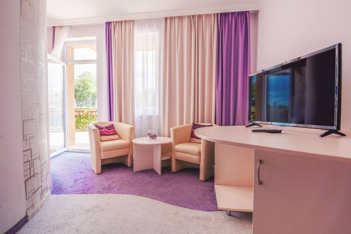 Habitación de hotel con una televisión grande, dos sillones, una mesita y vistas al exterior a través de cortinas traslúcidas.