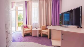 A hotel room with a large TV, two armchairs, a small table, and a view of the outdoors through sheer curtains.
