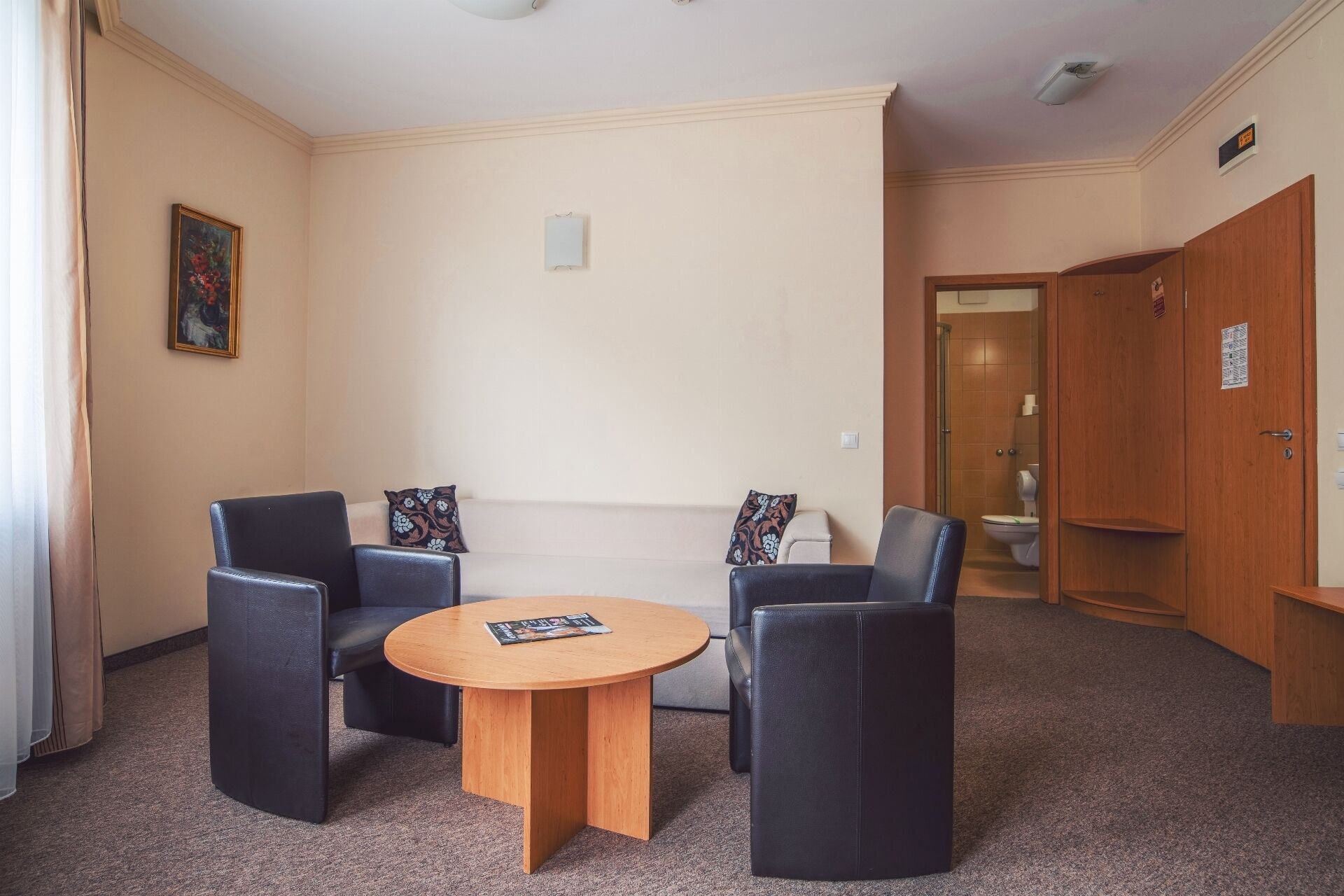 Ein Zimmer mit einem runden Holztisch, zwei schwarzen Ohrensesseln und einem beige farbenen Sofa. An der Wand hängt ein Gemälde und eine Tür führt ins Badezimmer.