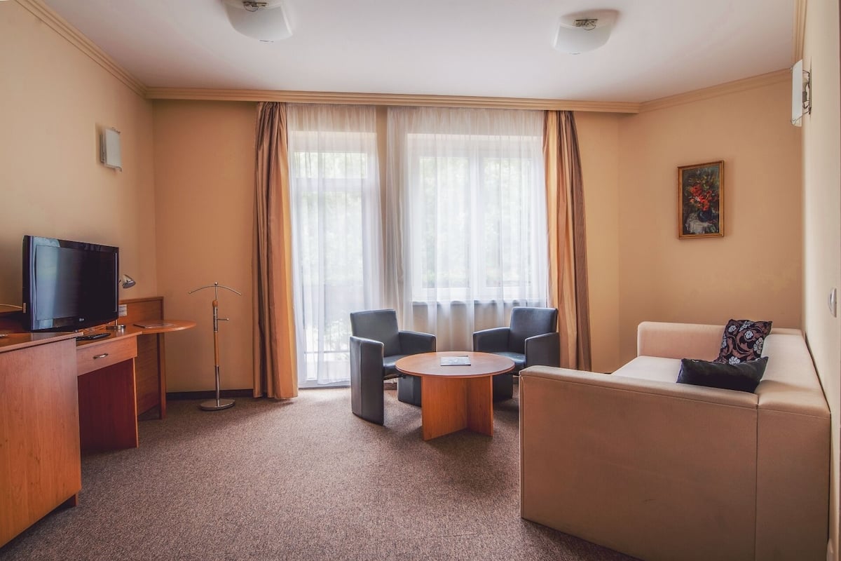 Habitación de hotel con televisión, escritorio, silla, sofá y ventana con cortinas.