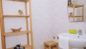 A bathroom with a wooden shelf unit, a small wooden stool, a white sink with a yellow basket, and a mirror above the sink. - Piccolo Sogno Inn (Pompei)