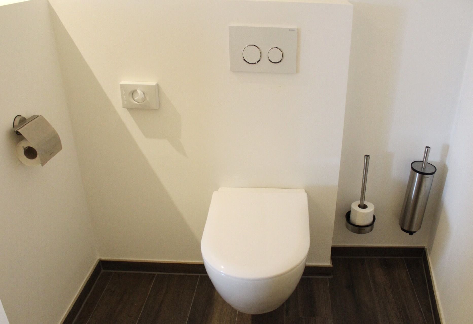 Un baño pequeño y moderno con inodoro blanco, dispensador de papel higiénico, bidé y pulsador de descarga en la pared.