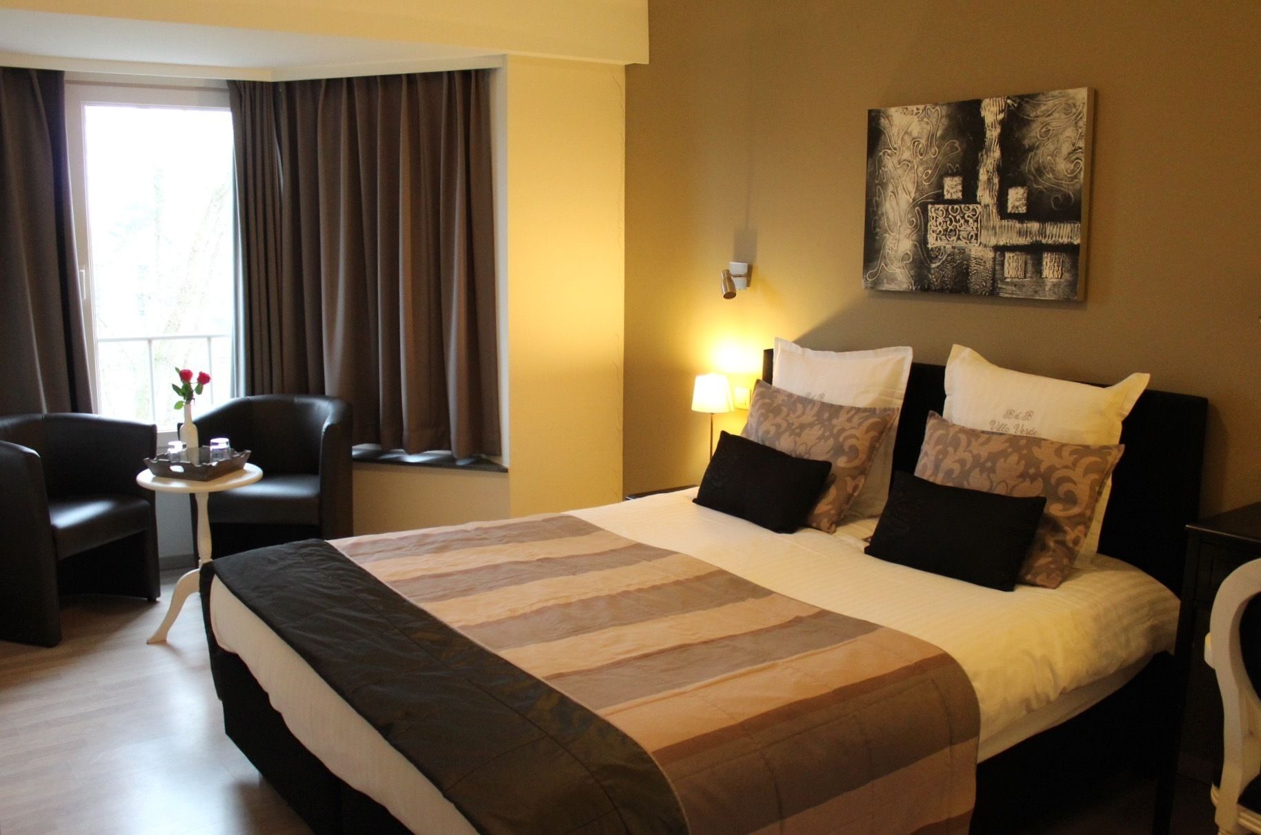 Een hotelkamer met een groot bed, twee stoelen, een kleine tafel met een bloemenvaas, en een schilderij aan de muur.